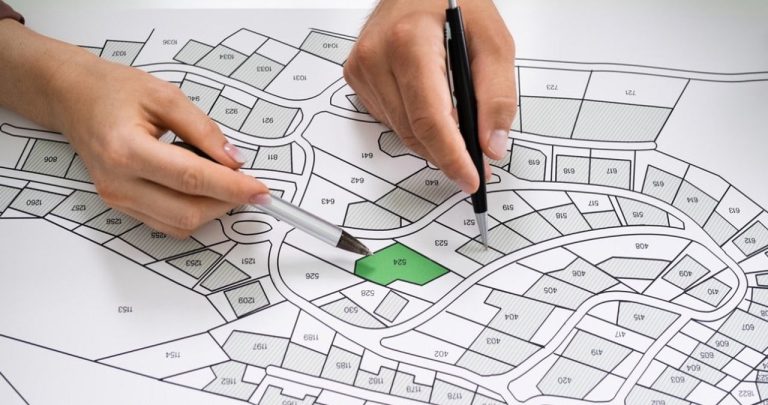 Un inginer topograf măsurând un teren rural cu un aparat GPS pe trepied, în timp ce proprietarul îi înmânează niște documente