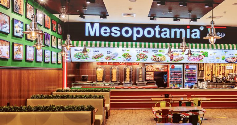 O tavă cu un meniu Kebab de la Mesopotamia pe o masă de food-court, alături de un bon fiscal și un smartphone deschis pe pagina de contact