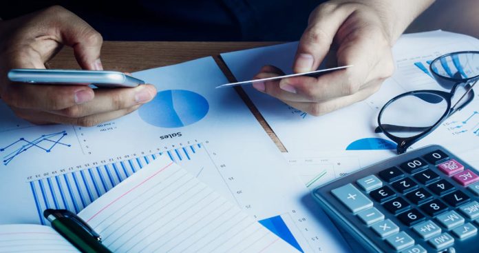 Un utilizator verificând grafice de trading pe telefon lângă un laptop cu Declarația Unică deschisă