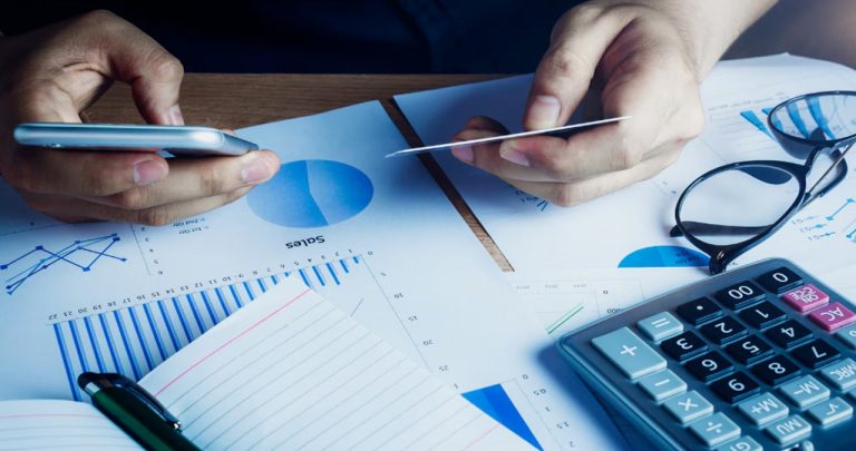 Un utilizator verificând grafice de trading pe telefon lângă un laptop cu Declarația Unică deschisă