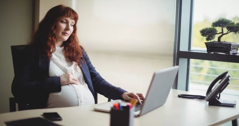 O femeie însărcinată zâmbind la birou în fața laptopului ținând în mână o adeverință medicală pentru departamentul HR